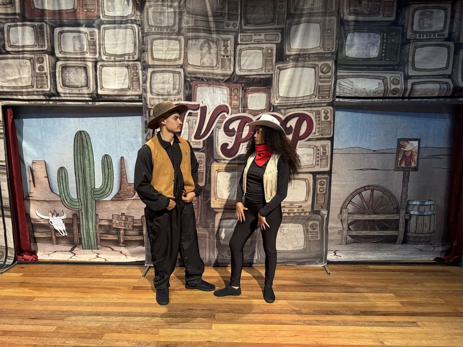 Um caubói e uma cowgirl se encaram diante de um cenário de deserto com cactos e placa Old West